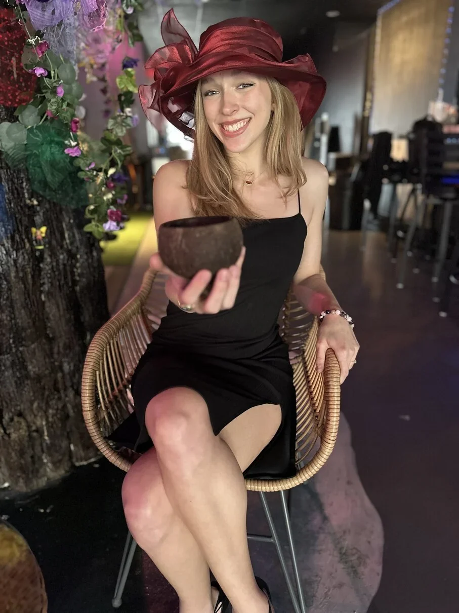 A smiling woman in a burgundy hat holds a coconut-shell cup while sitting inside Tribe Tea Lounge