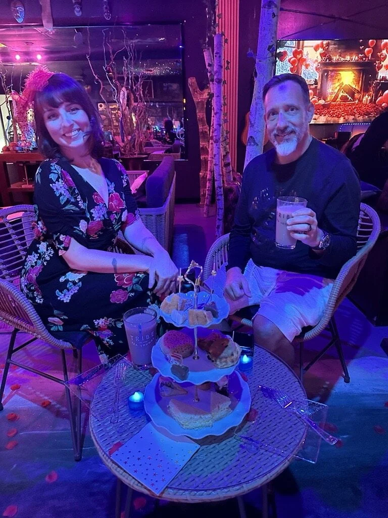 Two people enjoy drinks and a tiered tray of treats at a small table inside Tribe Tea Lounge