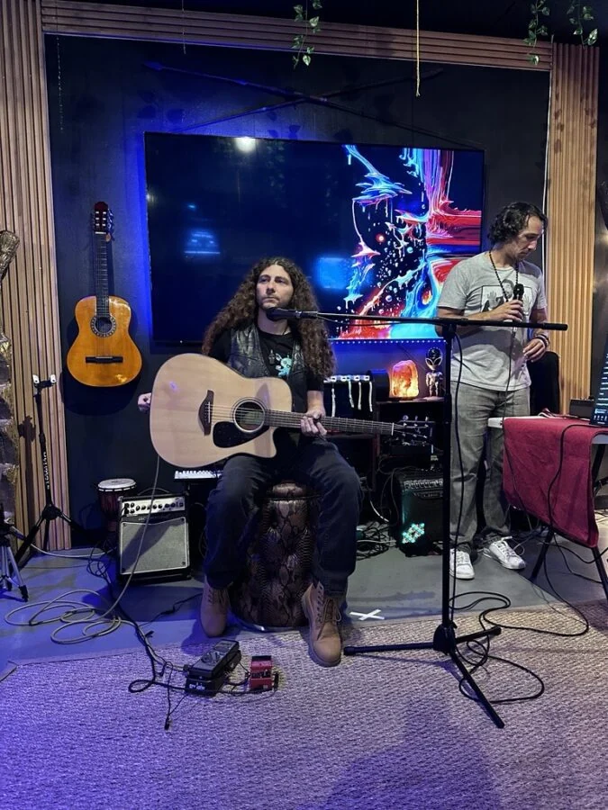 A seated guitarist and a standing musician perform with microphones and instruments at Tribe Tea Lounge