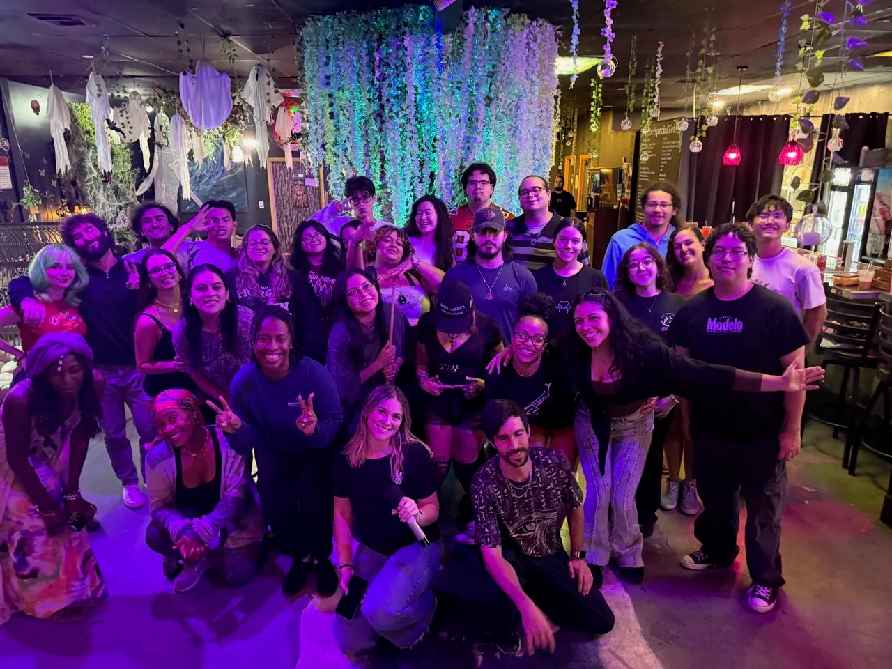 A large group smiles together beneath hanging flowers inside Tribe Tea Lounge