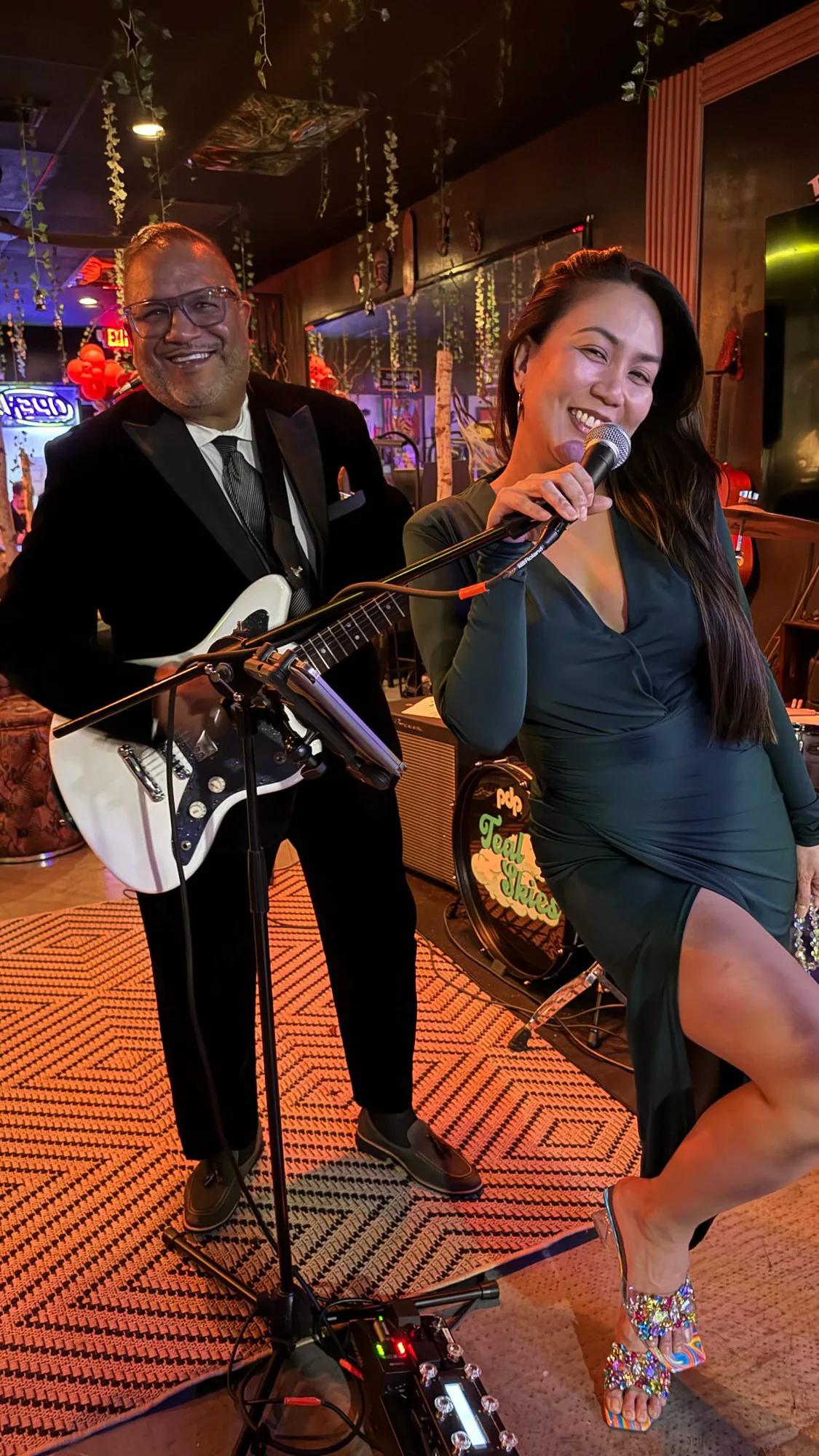 Tariq holds a white guitar beside Rose, who smiles with a microphone inside Tribe Tea Lounge