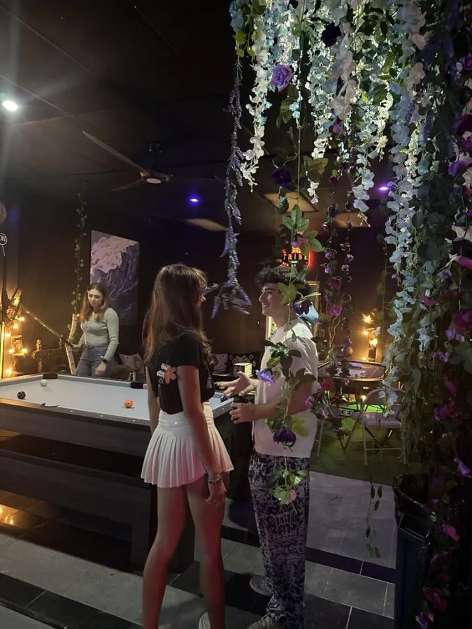 People talk beside a pool table beneath hanging purple flowers inside Tribe Tea Lounge