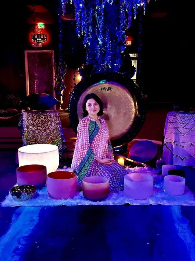 A woman sits behind a display of sound bowls and a large gong under Tribe Tea Lounge’s hanging flowers